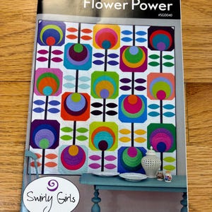 May include: A quilt pattern titled "Flower Power" featuring a colorful, geometric floral design. The quilt design includes squares with concentric circles in various colors, including orange, purple, green, and blue. The pattern is by Swirly Girls.