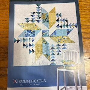 May include: A quilt pattern titled "Wind Drifter" by Robin Pickens. The pattern features a star design in shades of blue, yellow, and white. The image includes a chair with a stack of fabric and the text "Fat Eighth-Friendly Pattern in Two Sizes."