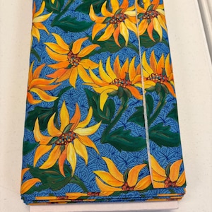 May include: Folded fabric with a vibrant floral pattern. The design features large yellow and orange sunflowers with green leaves against a blue background with a swirling pattern. The fabric is folded neatly, showing the repeating design.