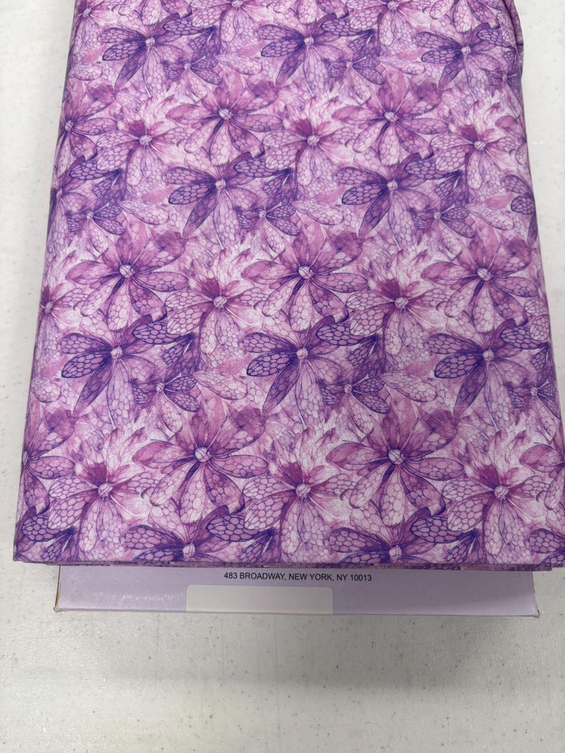 K&ouml;nnte beinhalten: Gefalteter Stoff mit einem sich wiederholenden Blumenmuster in Lila- und Wei&szlig;t&ouml;nen. Die Blumen haben ein detailliertes, netzartiges Design. Der Stoff liegt auf einer hellvioletten Schachtel. Der Text "483 BROADWAY, NEW YORK, NY 10013" ist auf der Schachtel zu sehen.