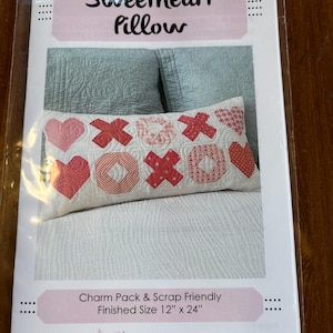 May include: A pillow pattern packet titled "Sweetheart Pillow." The pillow features a design of hearts and X's and O's in red and white. Finished size is 12" x 24". The packet is charm pack and scrap friendly.
