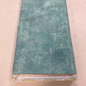 May include: A teal blue fabric with a white grid pattern and a subtle distressed look. The fabric has a Robert Kaufman label with the text "Robert Kaufman Fabrics" and a website address.