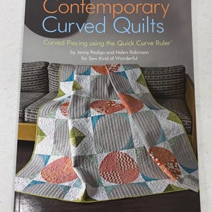 May include: A book titled "Contemporary Curved Quilts" featuring a colorful quilt with curved designs. The quilt has gray, orange, blue, and green circular and geometric patterns. The book cover also displays the text "Curved Piecing using the Quick Curve Ruler".