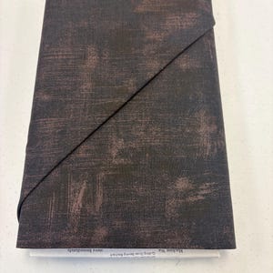 May include: Folded fabric with a dark brown and rust-colored distressed pattern. The fabric is folded over with a black elastic band. The text "Machine Wa... move immediately." is visible on a white label.