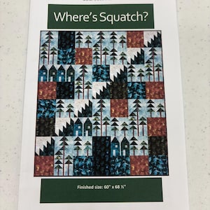 May include: A printed quilt pattern titled "Where's Squatch?" features a patchwork design with houses, trees, and geometric shapes in shades of blue, brown, and white. The finished size is 60" x 68 ½".