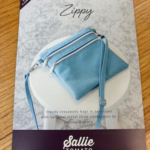 May include: A sewing pattern for a crossbody bag called "Zippy" by Sallie Tomato. The pattern includes instructions for making two sizes of bags with optional metal strap connectors. The image shows a light blue bag with white zippers.