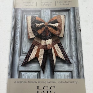 May include: A sewing pattern for a quilted bow, with the title "The Big Giant Quilted Jelly Roll Bow". The bow is made from fabric strips in brown, beige, and black. The pattern is designed for 2.5" strips and is reversible. The text "LGC" is also visible.