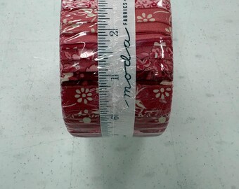 May include: A roll of red fabric with a floral pattern, wrapped in clear plastic. A white measuring tape with the word "moda" and the text "FABRICS + SUPPLIES" is wrapped around the fabric roll.