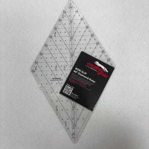 May include: A clear, diamond-shaped quilting ruler with grid lines and measurements, accompanied by a black and red product label. The label reads "NON-SLIP 60° Diamond Ruler" and "Made in USA".