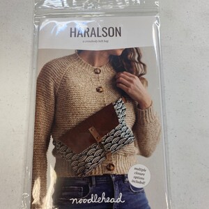 May include: A sewing pattern for a crossbody belt bag called "Haralson" by Noodlehead. The pattern includes multiple closure options. The image shows a woman wearing a brown sweater and jeans with the bag.