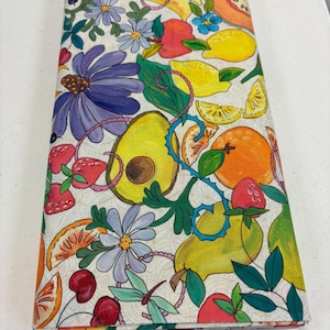 May include: A folded fabric with a colorful fruit and floral pattern. The design includes avocados, oranges, lemons, strawberries, cherries, and bananas, along with blue and purple flowers. The fabric has a cream-colored background.