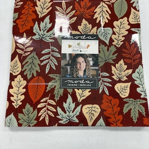 May include: A stack of square fabric pieces featuring a repeating pattern of autumn leaves in shades of dark red, orange, cream, and sage green. A paper label on the front displays the text "moda Windfall" and "moda FABRICS + SUPPLIES".