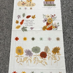 May include: A white fabric panel featuring autumn-themed illustrations including pumpkins, a stack of books with the text between the pages of a book is the COZIEST place to be, falling leaves with text, a coffee mug, and various sunflowers.