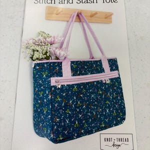 May include: A Stitch and Stash Tote pattern cover featuring a quilted teal bag with a colorful scissor print, light purple handles, and a zippered front pocket. The bag is shown hanging on a wooden hook with pink flowers inside. Knot + Thread Design.