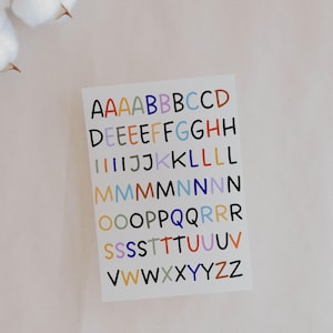 Könnte beinhalten: Ein weißes Aufkleberblatt mit bunten Großbuchstaben. Die Buchstaben sind in verschiedenen Farben, darunter Rot, Blau, Grün und Orange. Die Buchstaben sind in Reihen und Spalten angeordnet, mit mehreren Vorkommnissen jedes Buchstabens.