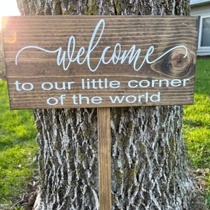 May include: A wooden sign with the text "Welcome to our little corner of the world" in white lettering. The sign is attached to a wooden stake.