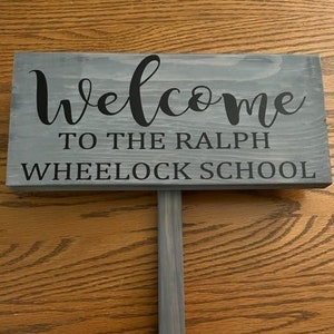 May include: A gray wooden sign with black text that reads "Welcome to the Ralph Wheelock School".