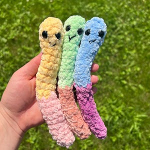 Puede incluir: Tres coloridos gusanos de ganchillo hechos a mano con caras sonrientes. Los gusanos son amarillos, verdes y azules, con fondos rosas, naranjas y morados. Los juguetes de peluche blandos se sostienen contra un fondo verde borroso.