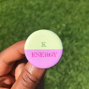 Op de afbeelding: Een ronde knop met een groene bovenhelft en een paarse onderhelft. De bovenhelft heeft een groene letter "K" en de onderhelft heeft het woord "ENERGY" in roze.