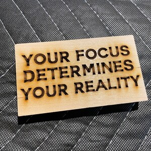 May include: A rectangular wooden plaque with the engraved text "YOUR FOCUS DETERMINES YOUR REALITY". The text is in a bold, sans-serif font, and the wood has a light brown colour. The plaque is set against a black quilted background.