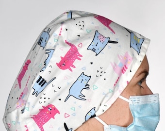 May include: A white scrub cap with a colorful pattern of cats and geometric shapes. The cap is worn by a person wearing a blue scrub top and a light blue surgical mask.