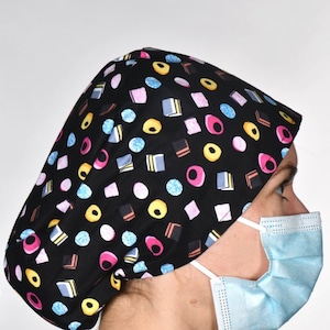 Peut inclure: Une coiffe chirurgicale noire ornée d'un motif de bonbons colorés, portée avec un masque chirurgical bleu. Le bonnet présente diverses formes et couleurs, notamment rose, jaune, bleu et blanc. La personne porte un haut de travail bleu.