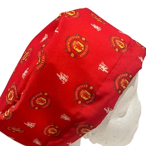 May include: Red fabric cap featuring a repeating pattern of the Manchester United crest and logo. The crest is a shield with a ship and the words "Manchester United" in a circular border. The cap is made of a soft, flexible material.