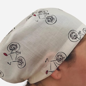 May include: A white cap featuring a repeating pattern of black bicycles with red details. The cap is made from a soft material and is designed to fit the head. The bicycle design gives a playful look.