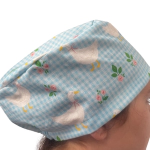 May include: A light blue and white gingham scrub cap featuring a repeating pattern of white geese with yellow feet and small pink rose clusters with green leaves. The fabric cap has a rounded crown and a wide band that fits around the head.