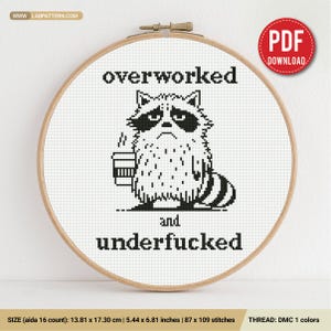 Puede incluir: Un patrón de punto de cruz en un bastidor de madera con un mapache pixelado negro sosteniendo una taza de café. El texto dice overworked and underfucked. El diseño mide 13,81 por 17,30 cm en tela Aida de 16 hilos, usando un solo color.