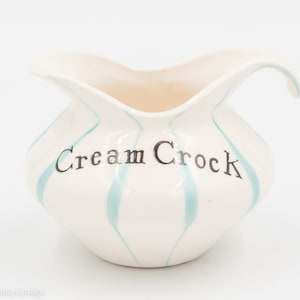 May include: White ceramic creamer with a blue and white striped design. The creamer has a handle and says "Cream Crock" on the front.