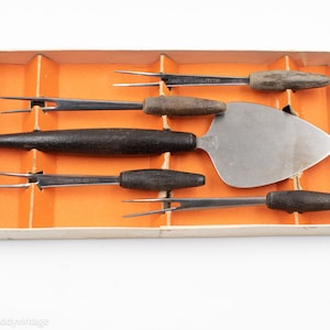 May include: A set of five vintage silver-toned cheese knives with wooden handles in a cardboard box. The knives include a spreader, a fork, and three small forks.