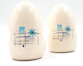 May include: A pair of vintage ceramic salt and pepper shakers. The shakers are egg-shaped with a speckled cream-colored finish. Each shaker features a mid-century modern design with blue and gray geometric shapes and a black grid pattern.