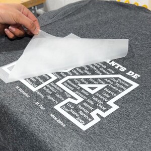 May include: A gray t-shirt with a white graphic design featuring the number 4 and a list of names. A hand is peeling away a transparent transfer sheet, revealing the design.