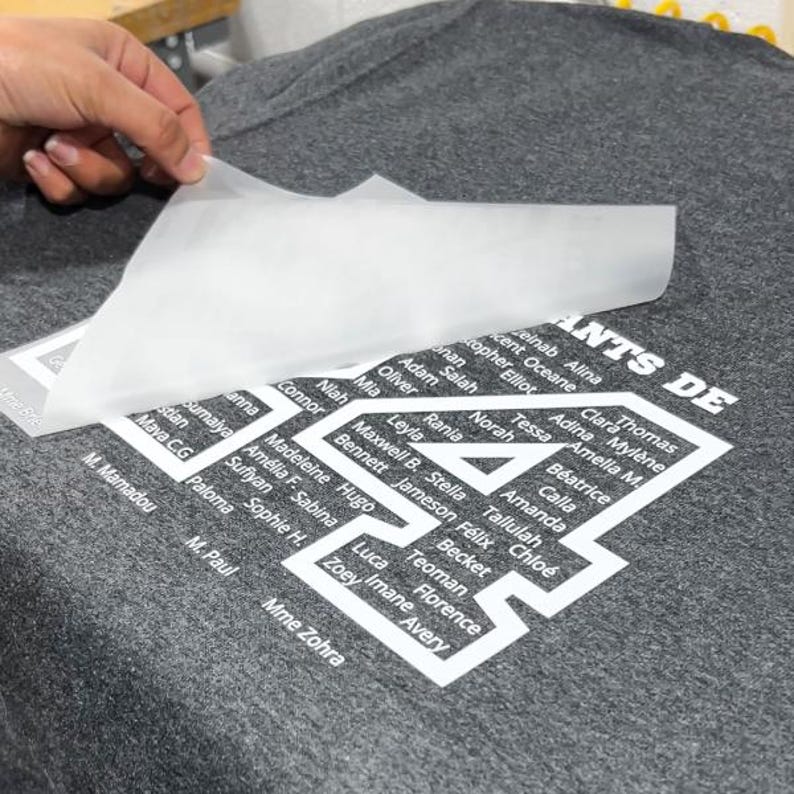 May include: A gray t-shirt with a white graphic design featuring the number 4 and a list of names. A hand is peeling away a transparent transfer sheet, revealing the design.