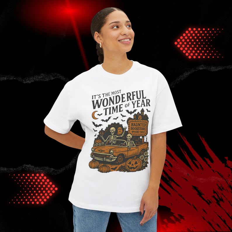 May include: White t-shirt with a Halloween-themed graphic. The design features a vintage orange car with skeletons, pumpkins, bats, and the text "It's the Most Wonderful Time of Year." The car is in front of a haunted bookstore.