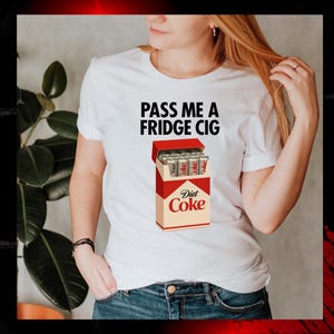 K&ouml;nnte beinhalten: Wei&szlig;es T-Shirt mit dem Text "PASS ME A FRIDGE CIG" und einer Grafik einer Diet Coke Zigarettenschachtel. Die Schachtel ist rot und beige, mit "Diet Coke" aufgedruckt. Das T-Shirt wird von einer Person getragen.