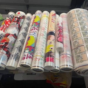 May include: A collection of rolled-up printed materials, featuring various colorful designs and patterns. Some rolls display cartoon characters, while others showcase geometric and floral motifs. A large roll of printed material is also visible.