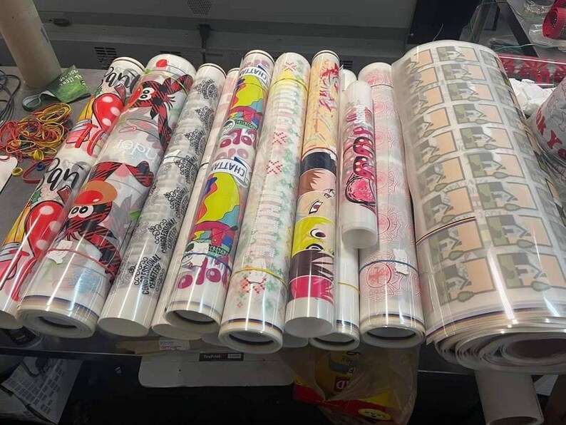 May include: A collection of rolled-up printed materials, featuring various colorful designs and patterns. Some rolls display cartoon characters, while others showcase geometric and floral motifs. A large roll of printed material is also visible.