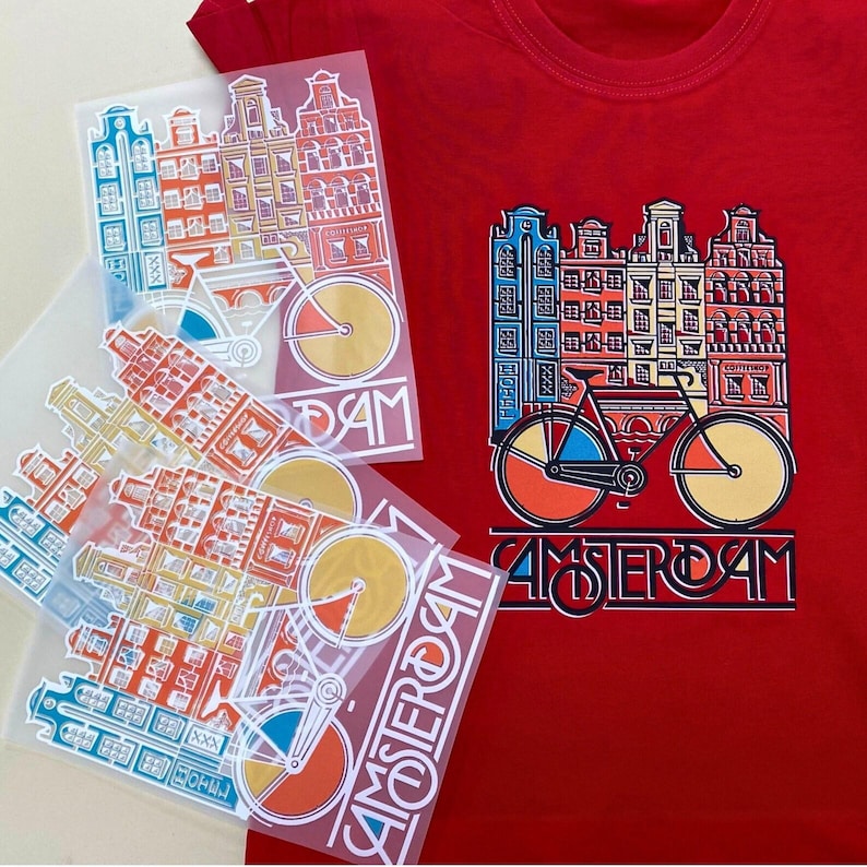 May include: Red t-shirt with a colorful graphic of Amsterdam buildings and a bicycle. The word "AMSTERDAM" is printed below the image. Several similar graphic transfers are also visible.