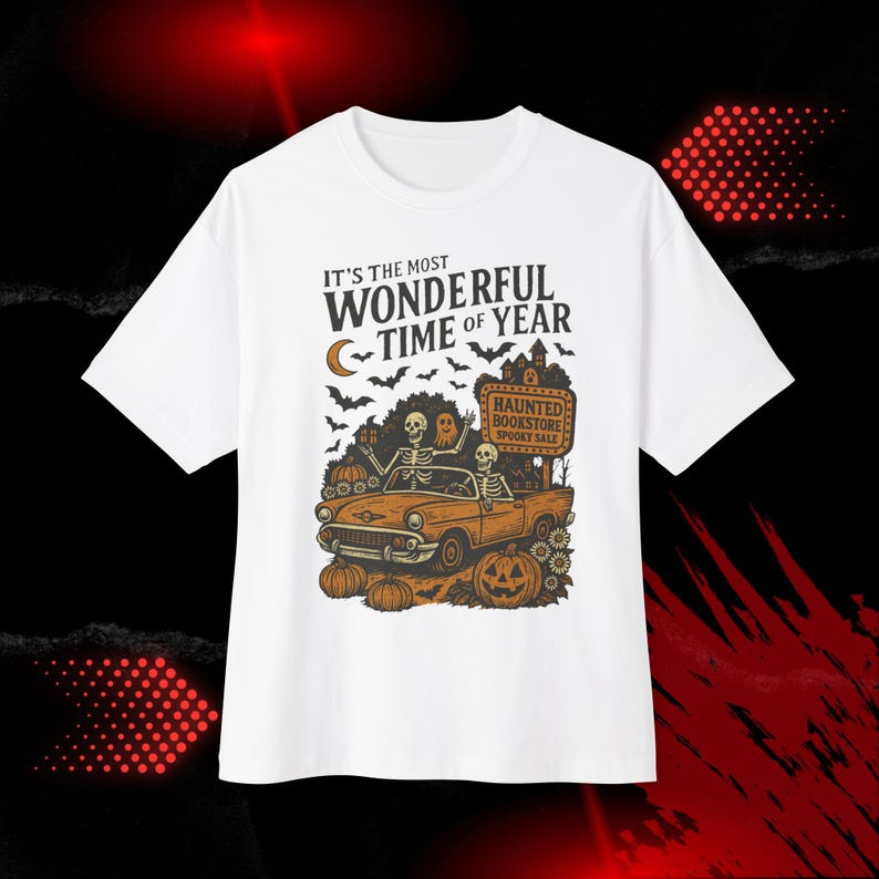 May include: White t-shirt with a Halloween-themed graphic. The design features skeletons in a vintage car, pumpkins, bats, and the text "IT'S THE MOST WONDERFUL TIME OF YEAR." A sign reads "HAUNTED BOOKSTORE SPOOKY SALE."