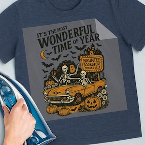 May include: Navy blue t-shirt with a Halloween-themed graphic. The design features skeletons in a vintage car, pumpkins, bats, and the text "It's the Most Wonderful Time of Year." A sign reads "Haunted Bookstore Spooky Sale."