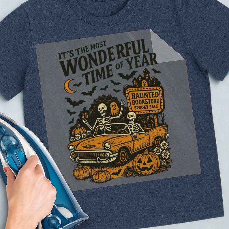 May include: Navy blue t-shirt with a Halloween-themed graphic. The design features skeletons in a vintage car, pumpkins, bats, and the text "It's the Most Wonderful Time of Year." A sign reads "Haunted Bookstore Spooky Sale."