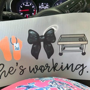 May include: A light gray sign with orange footprints, a black bow, and a gurney illustration. The text "he's working" is written in cursive. The sign is in front of a car's dashboard.