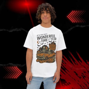 May include: White t-shirt with a Halloween-themed graphic. The design features a vintage orange car with skeletons, pumpkins, bats, and the text "IT'S THE MOST WONDERFUL TIME OF YEAR." A sign reads "Haunted Bookstore Spooky Sale."