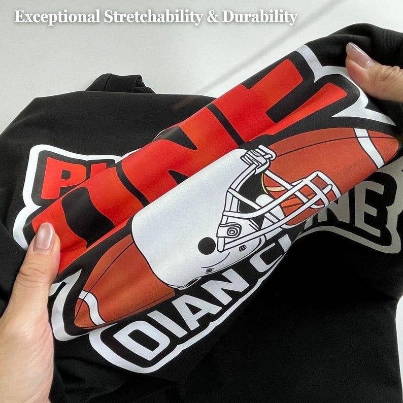 May include: Black sweatshirt with a football design. The design includes a football helmet and the words "INDIAN" and "PIONEERS" in white and red. The text "Exceptional Stretchability & Durability" is at the top.