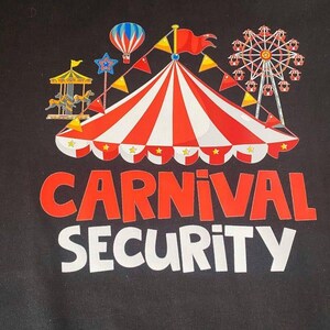 May include: Black t-shirt featuring a carnival design. The design includes a red and white striped circus tent, a Ferris wheel, a carousel, and the words "CARNIVAL SECURITY" in red and white lettering.