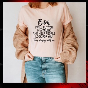 May include: A light pink t-shirt with the text "Bitch I will put you in a trunk and help people look for you. Stop playing with me" in black. The shirt is paired with blue jeans and a tan cardigan.