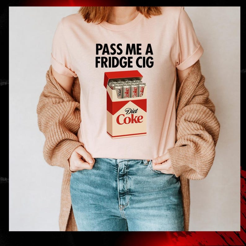 K&ouml;nnte beinhalten: Hellrosa T-Shirt mit dem schwarzen Text "PASS ME A FRIDGE CIG". Darunter befindet sich eine Grafik einer Diet Coke Zigarettenschachtel. Die Schachtel ist rot und beige, mit "Diet Coke" aufgedruckt.