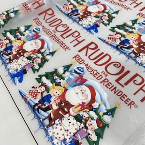 May include: Clear plastic sheet with a repeating pattern of Christmas characters, including Santa Claus, Rudolph the Red-Nosed Reindeer, and elves. The text "Rudolph the Red-Nosed Reindeer" is printed in red. The design features a festive holiday theme.